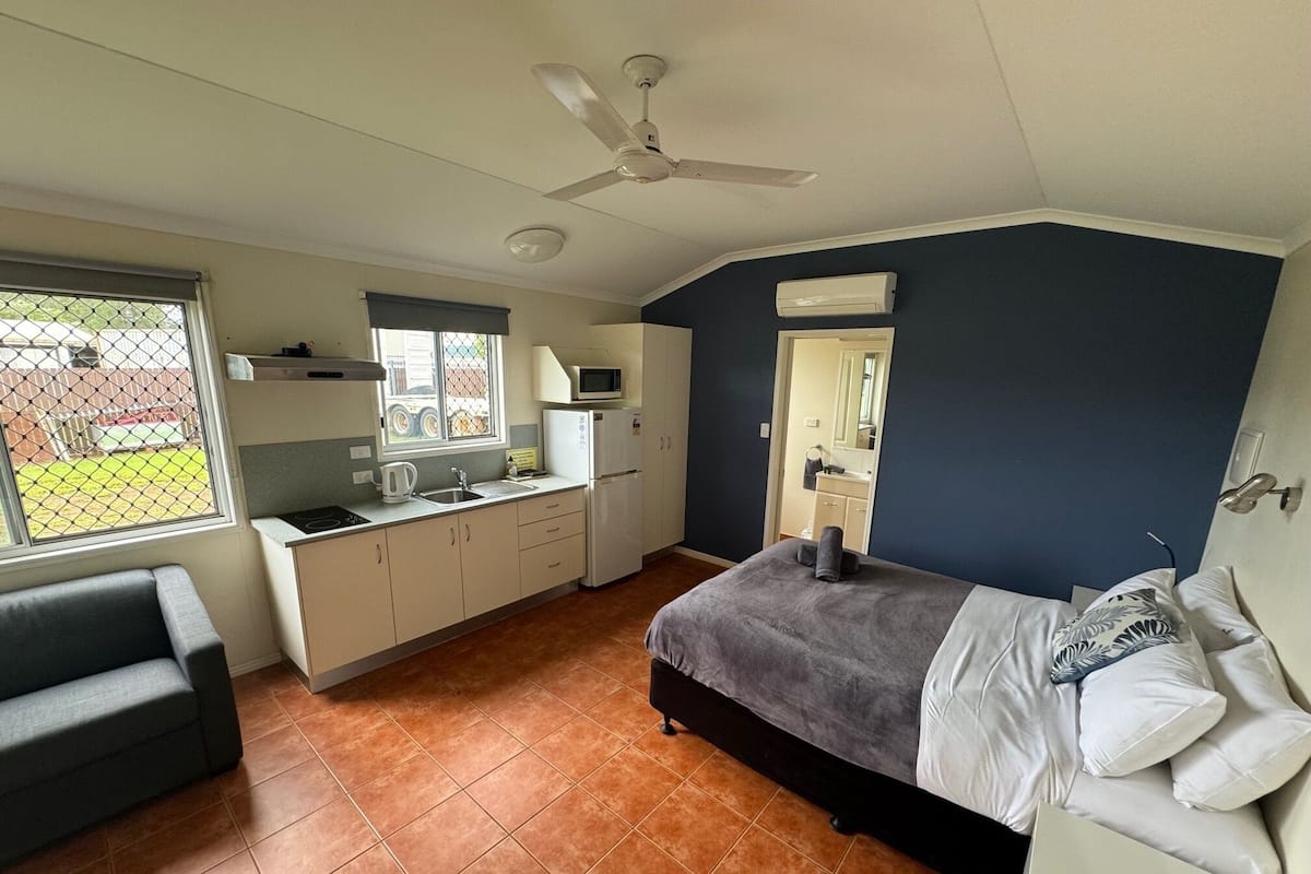 Une chambre compacte comprenant un lit, un petit coin cuisine, un ventilateur de plafond et une fenêtre donnant sur une courClose.