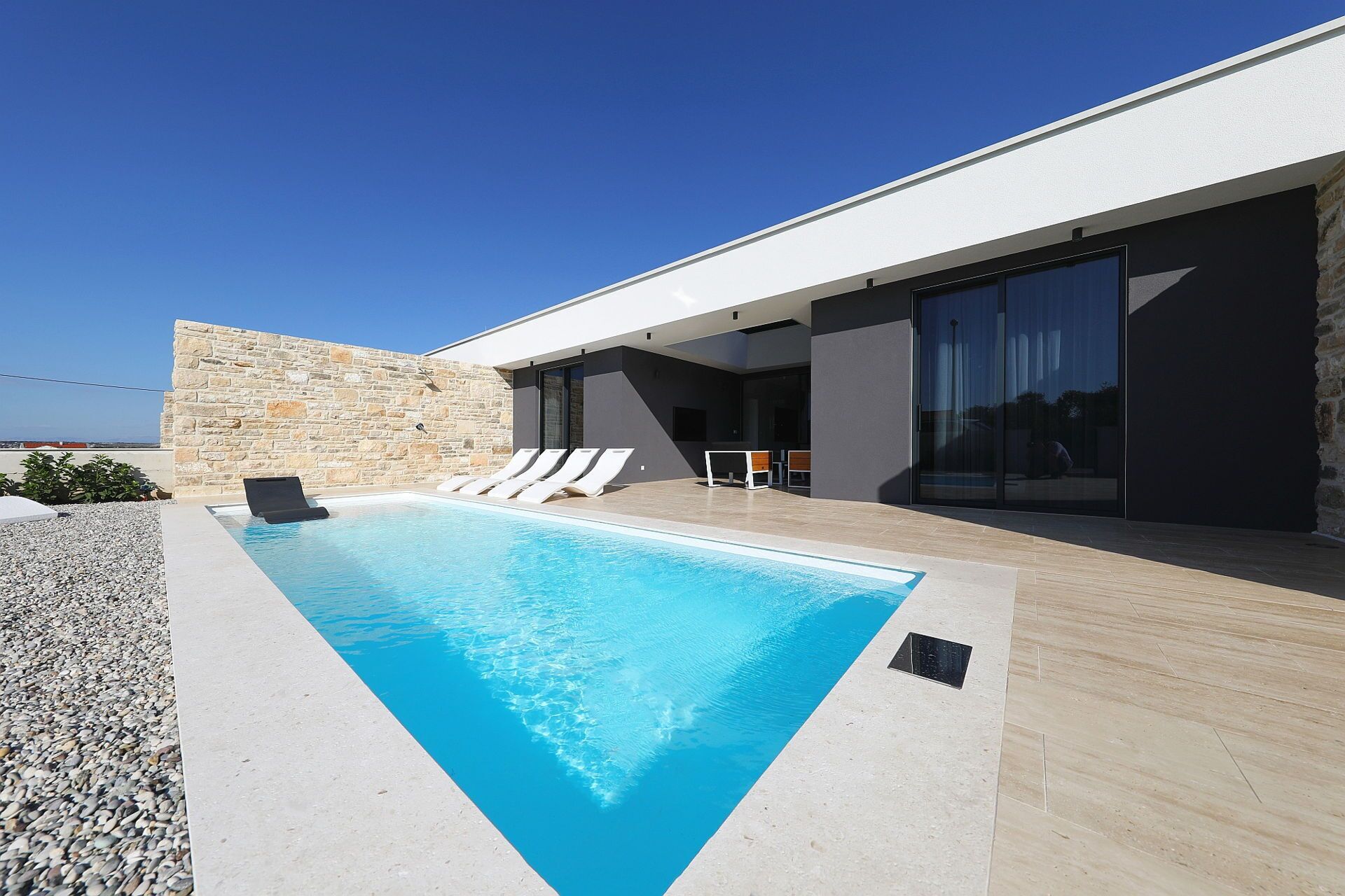 Un espace piscine moderne agrémenté d’un mur en pierre, d’une piscine et d’un coin salon couvert avec des chaises et une table.