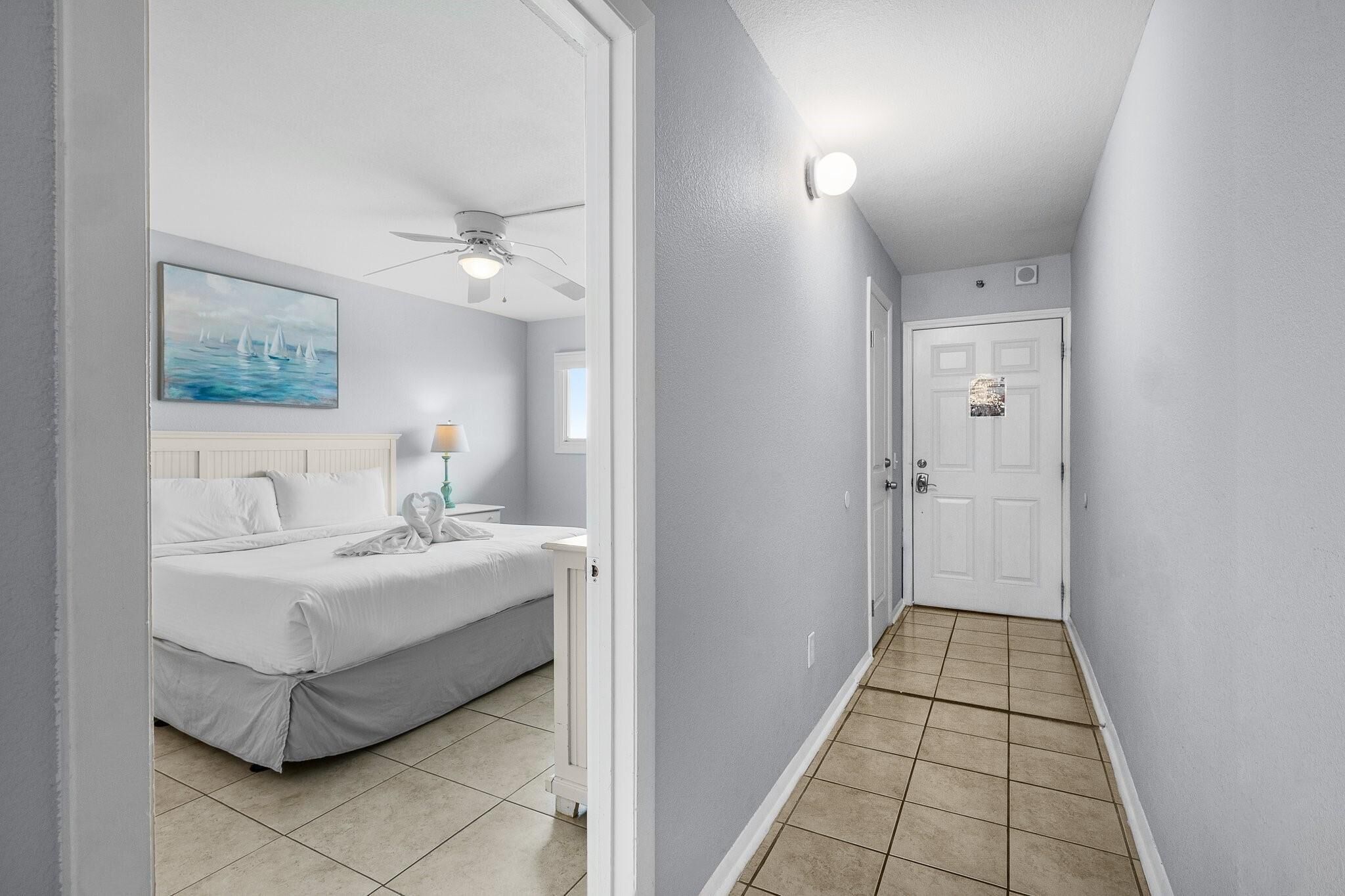 A bedroom with a bed, a ceiling fan, a painting, and a nightstand with a lamp.