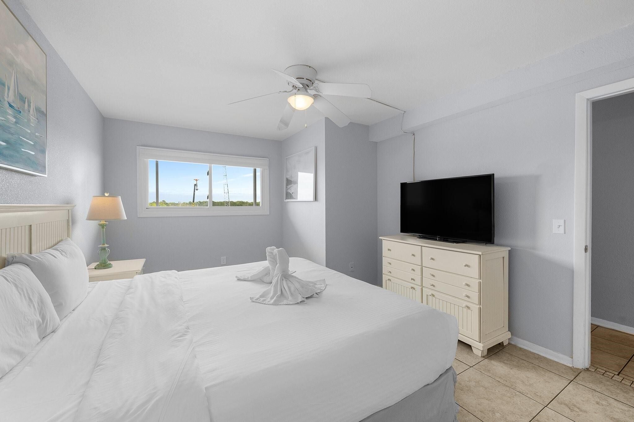 A neatly made bed with white linens, a television on a dresser, and a window with a view of the outdoors.