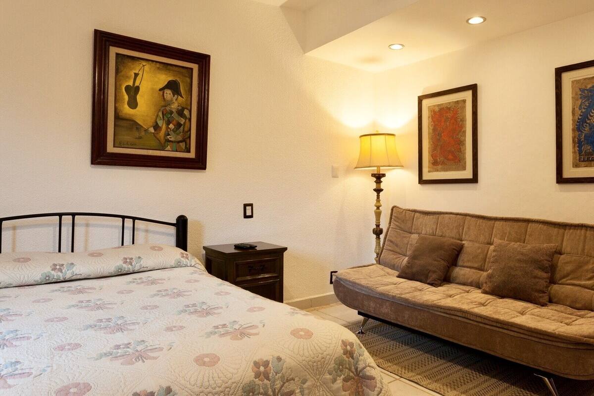 Un dormitorio con cama, sofá, mesita de noche, lámpara y cuadros en las paredes.
