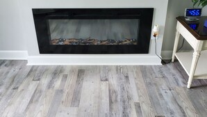 A room with a fireplace, wooden flooring, and a small table with a clock.