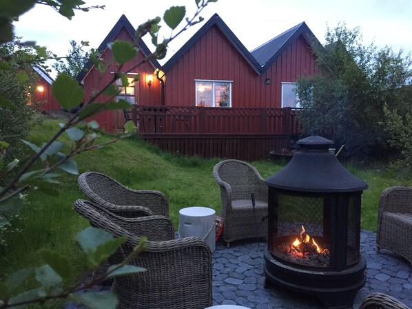 A cozy outdoor seating area with wicker chairs and a fire pit, set against a backdrop of red wooden cabins.