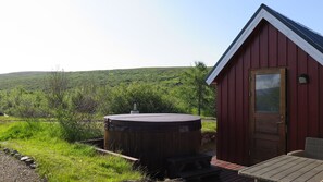 A wooden cabin with a hot tub and a deck, surrounded by lush greenery and hills.