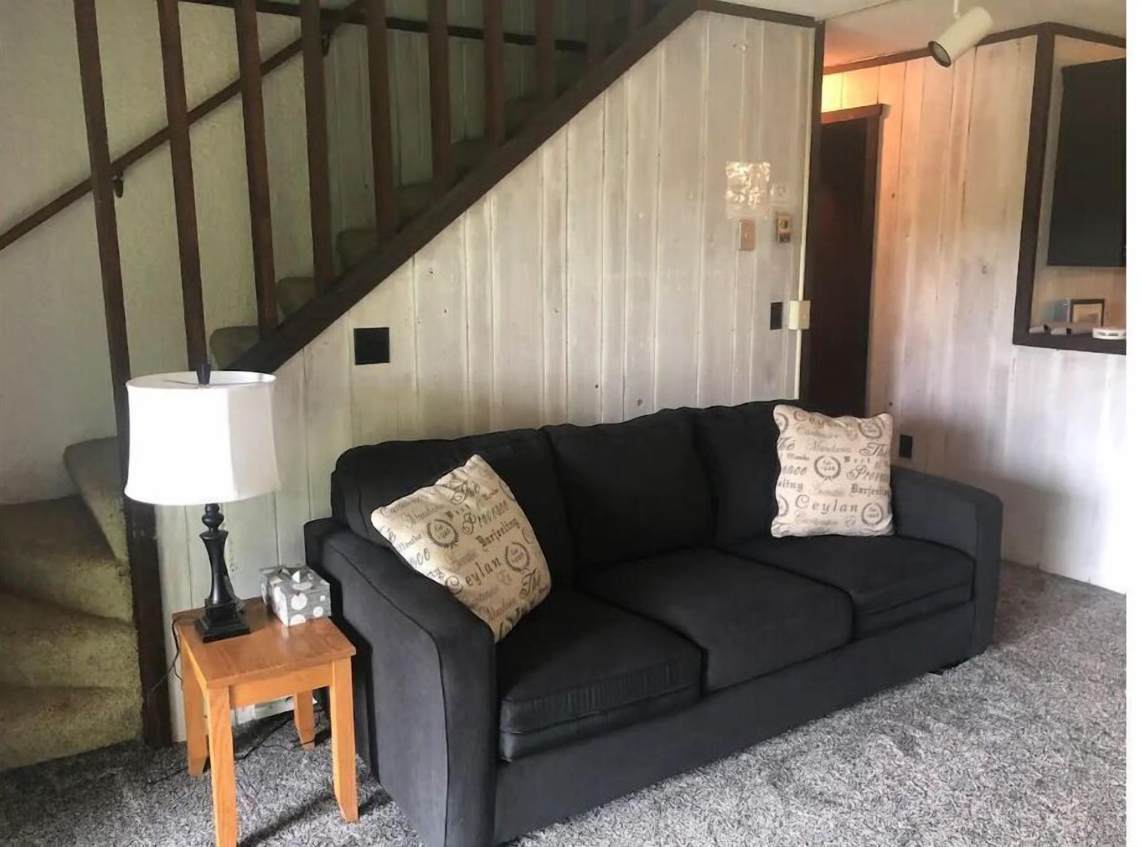 Un canapé noir agrémenté de coussins à motifs, une petite table d’appoint en bois avec une lampe, et un escalier en arrière-plan.