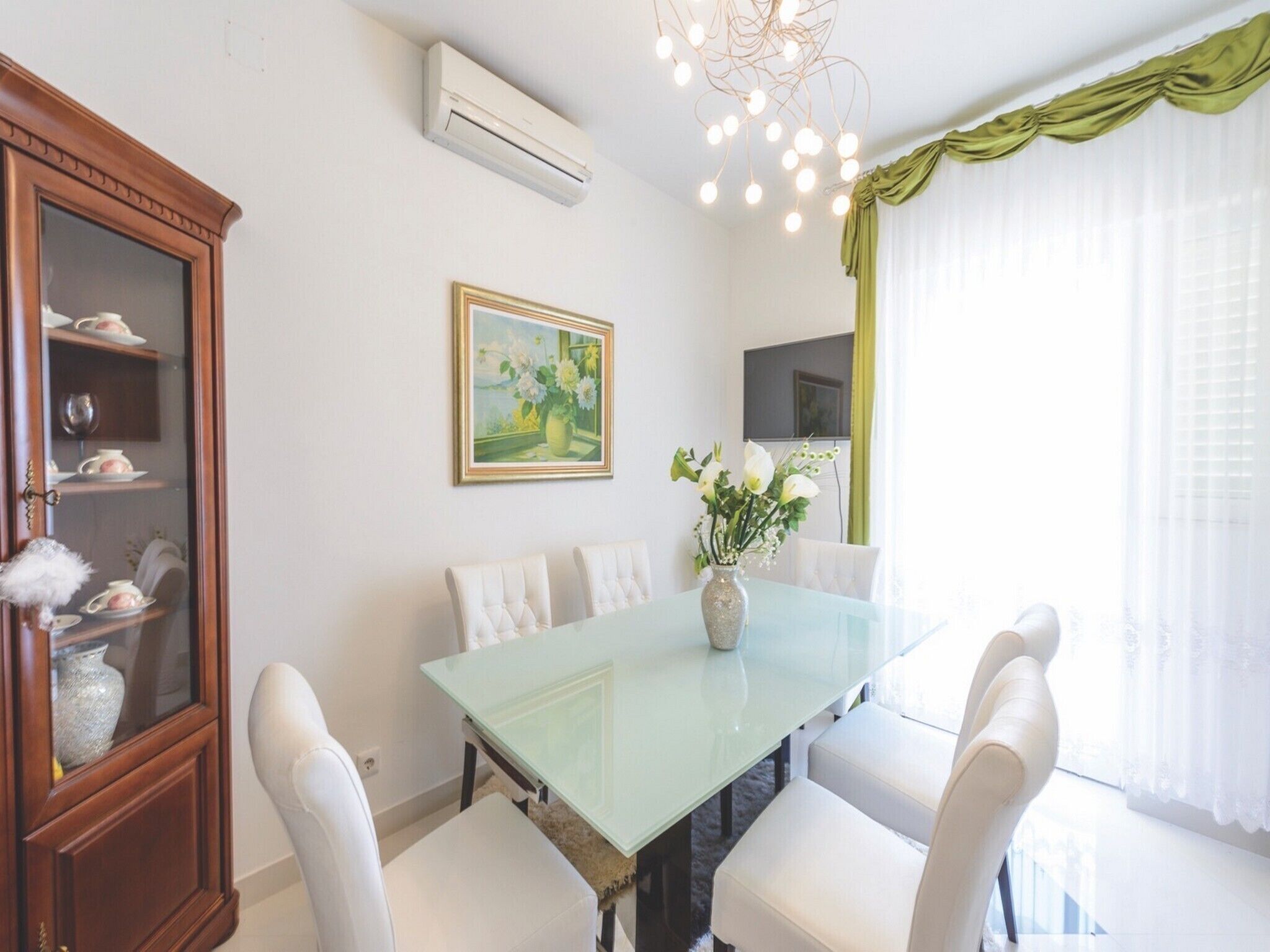 A dining area with a glass table, white chairs, a vase with flowers, a painting on the wall, and a green curtain.