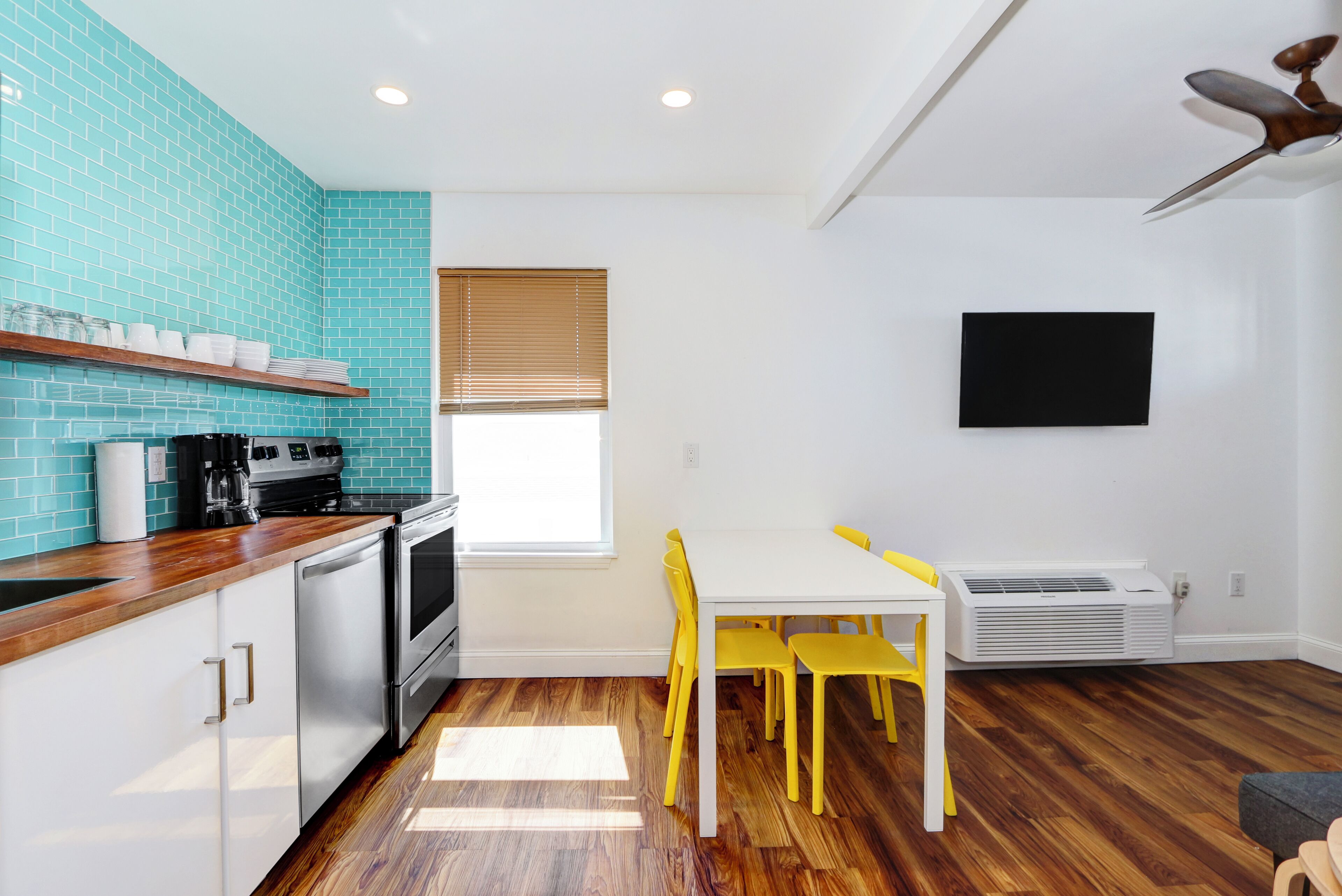 Una cocina con encimera de madera, armarios blancos y salpicadero de azulejos azules. Hay una mesa de comedor con sillas amarillas y un televisor blanco montado en la pared.