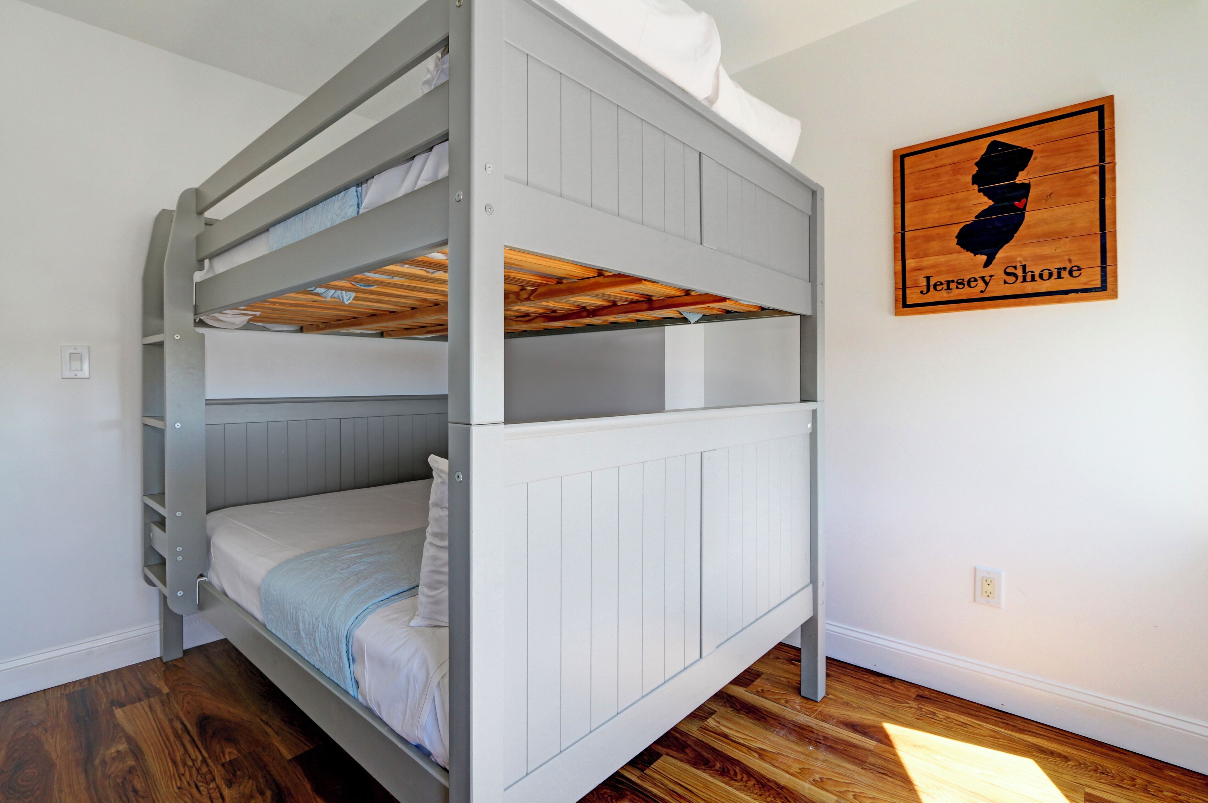 A bunk bed with white bedding and a wooden headboard. A framed map of Jersey Shore hangs on the wall.