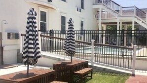 Un patio con bancos de madera y sombrillas a rayas, un sendero de ladrillo y un edificio blanco con un balcón al fondo.