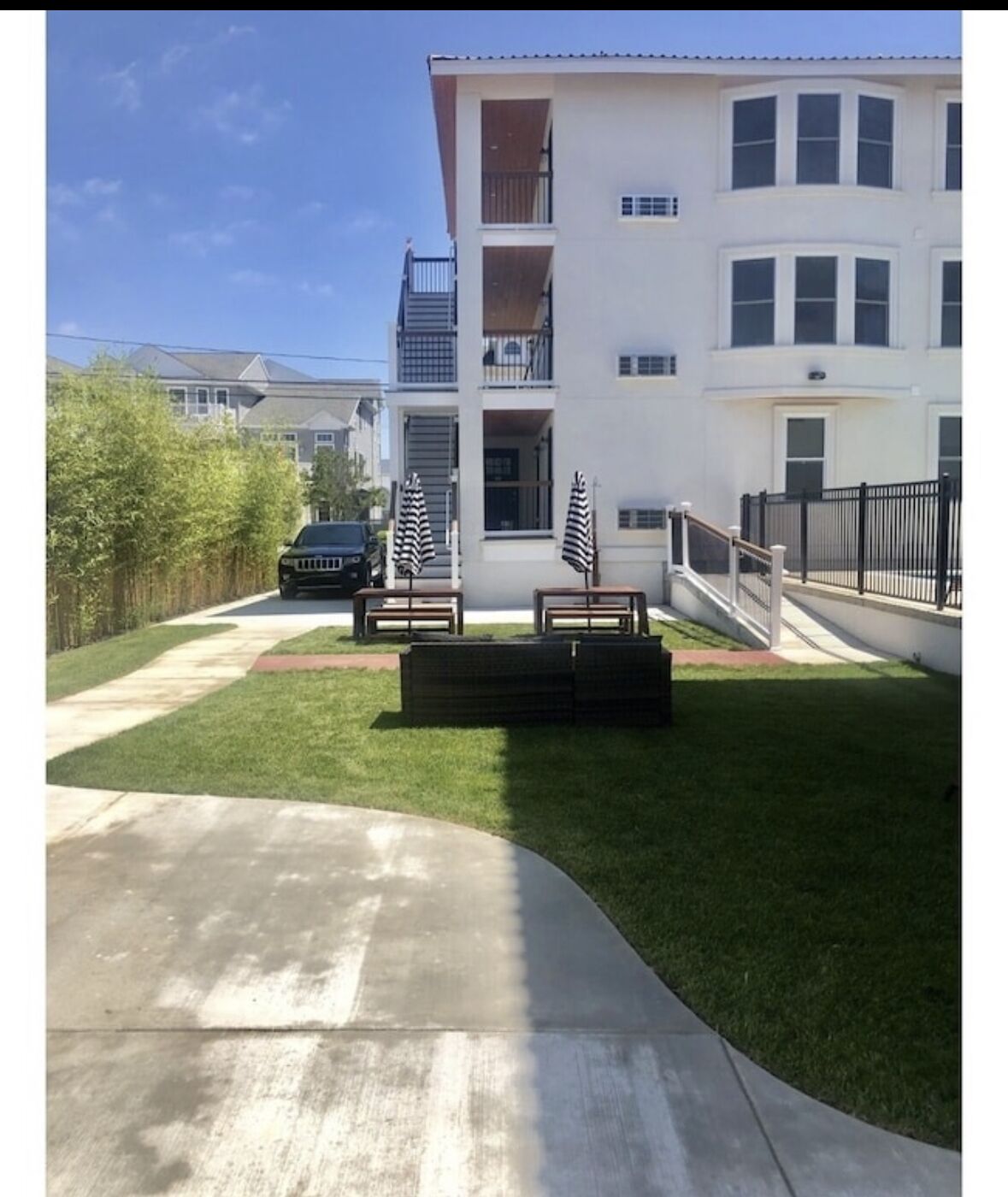Edificio residencial de dos plantas con un césped cuidado, zona de estar al aire libre con bancos y sombrillas, y un coche negro estacionado en la entrada.