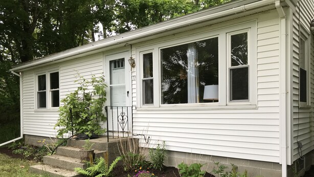 Einbungalow mit einer Veranda, ein kleiner Garten mit verschiedenen Pflanzen und Blumen sowie ein Metallzaun.