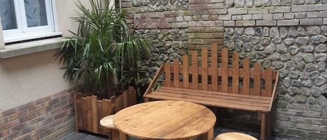 A wooden bench and table set on a deck with a stone wall and a window in the background.