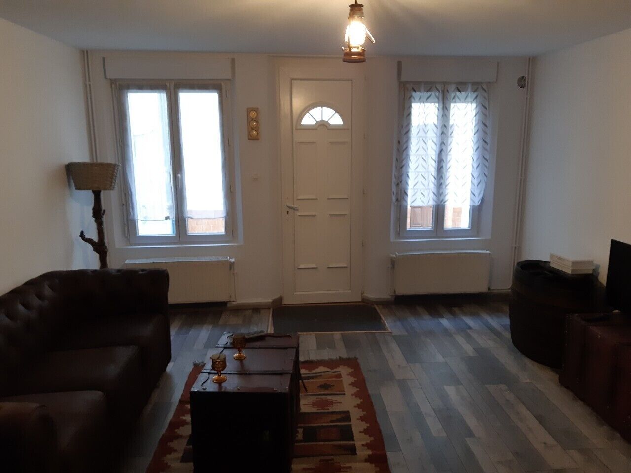 Una sala de estar con un sofá, una mesa de centro y una alfombra. Hay dos ventanas con cortinas, una puerta y una lámpara.