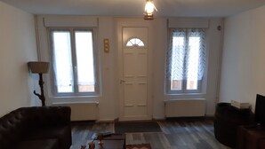 A living room with a sofa, a coffee table, and a rug. There are two windows with curtains, a door, and a lamp. - Le Cézard de Fleury (Le Tréport)