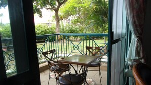 Un balcon avec une table et des chaises, une rambarde en métal vert et une vue sur un jardin avec des arbres et un bâtiment en arrière-plan.