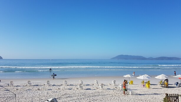 Ein Strand mit weißen Plastikstühlen und Sonnenschirmen, einem klaren blauen Himmel und ruhigen Wellen.