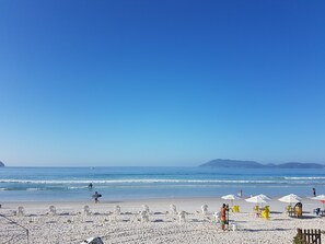 A beach with white plastic chairs and umbrellas, a clear blue sky, and calm sea waves.