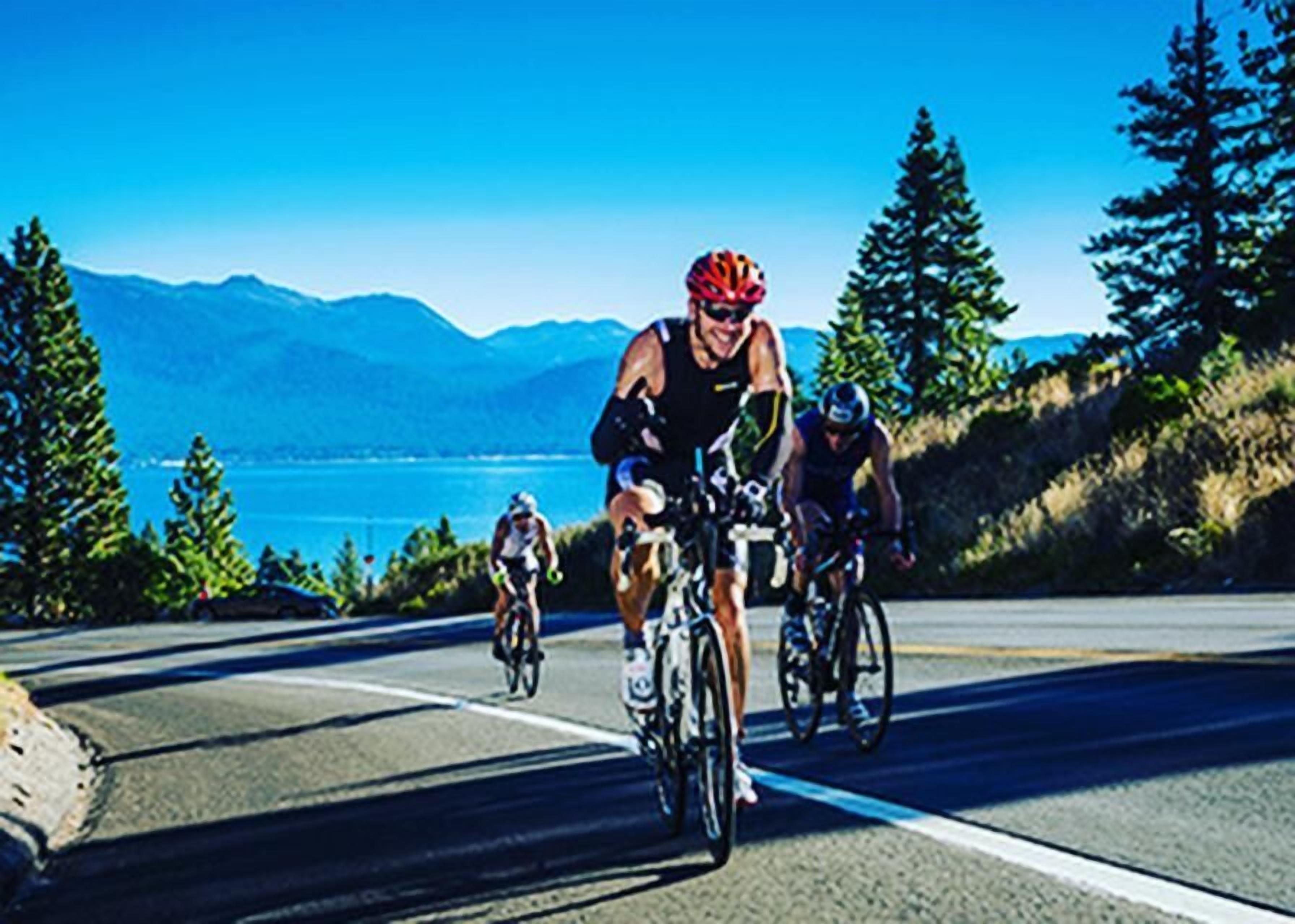 Três ciclistas em uma estrada com vista panorâmica de montanhas e um corpo d'água.