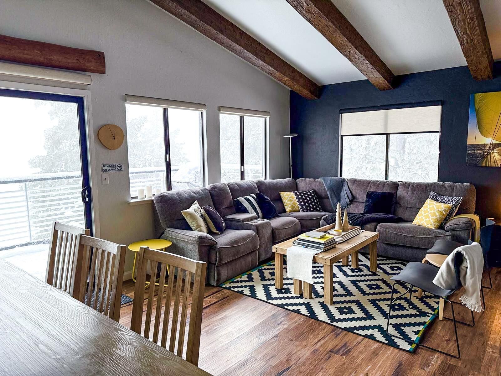 A cozy living room with a sectional sofa, wooden dining table, and chairs. There are large windows with blinds, a clock on the wall, and a rug on the floor.
