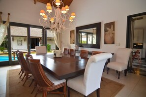 A dining area with a large wooden table, wicker chairs, and a white cushioned chair. A chandelier hangs above the table.