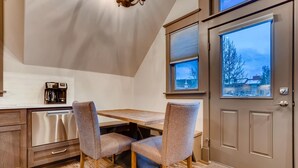 A kitchen with a dining table, two chairs, a coffee maker, and a dishwasher. There are two windows with blinds, and a door with a handle.