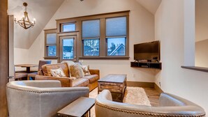 A cozy living room with a high ceiling, large windows, a chandelier, and a flat-screen TV.