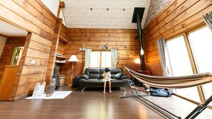 A log cabin interior with a wooden floor, a black leather sofa, a hammock, and a large window with curtains. - Loghouse Seseragi (Tsuru)