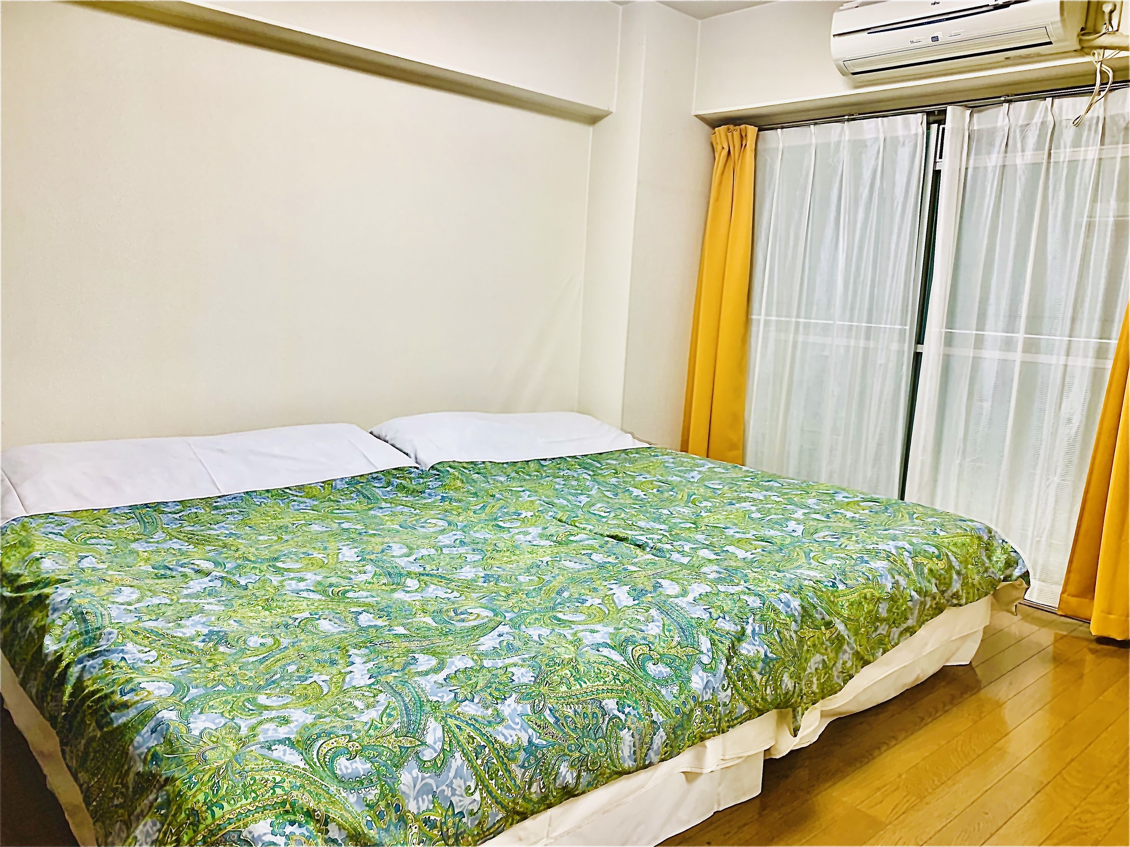 Une chambre à coucher avec un lit, des oreillers blancs, une couvre-lit à motifs verts, une fenêtre avec des rideaux jaunes et un appareil de climatisation placé au-dessus de la fenêtre.