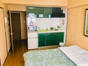 Una habitación pequeña con cama, kitchenette con microondas, lavamanos y un mueble verde.