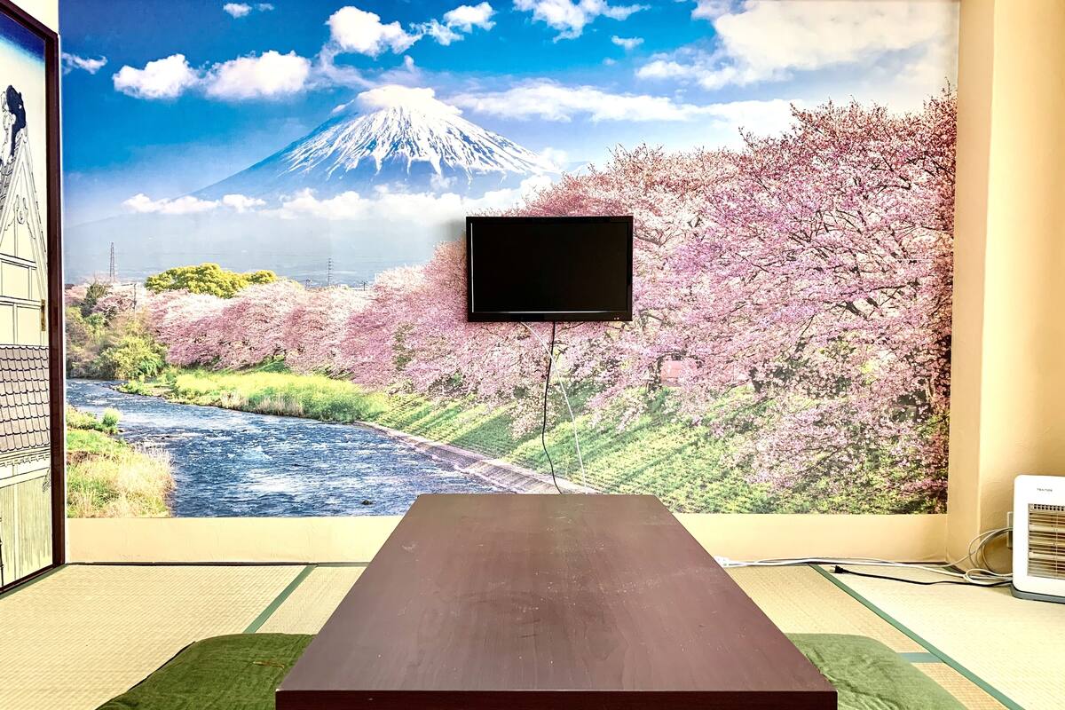 A room with tatami flooring, a wooden table, and a TV mounted on a wall displaying a scenic wallpaper of Mount Fuji and cherry blossoms.