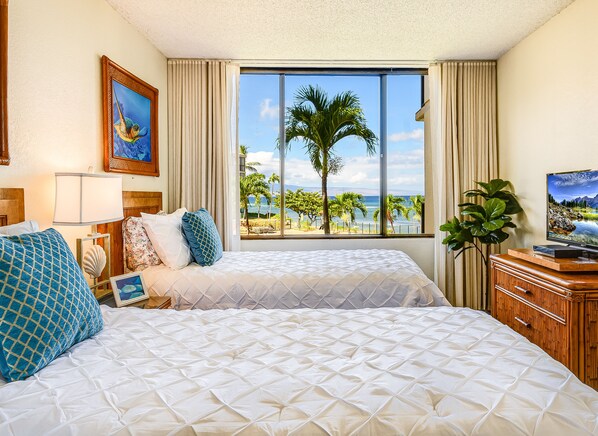 A hotel room with two beds, a large window with a view of palm trees and the ocean, a television on a wooden dresser, and a framed picture on the wall.