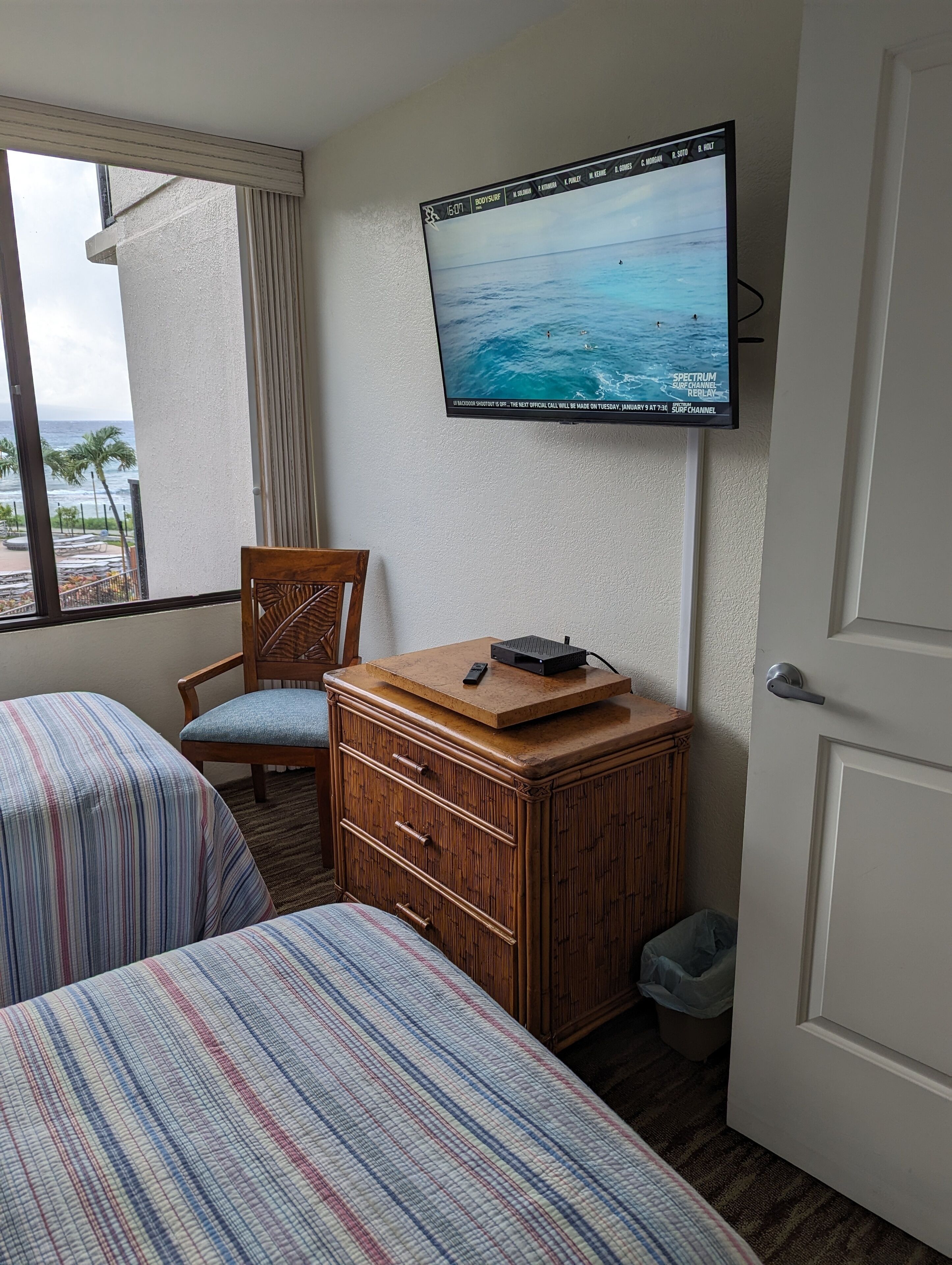 Habitación de hotel con dos camas, un aparador de madera, un televisor montado en la pared y vista al océano a través de una ventana.
