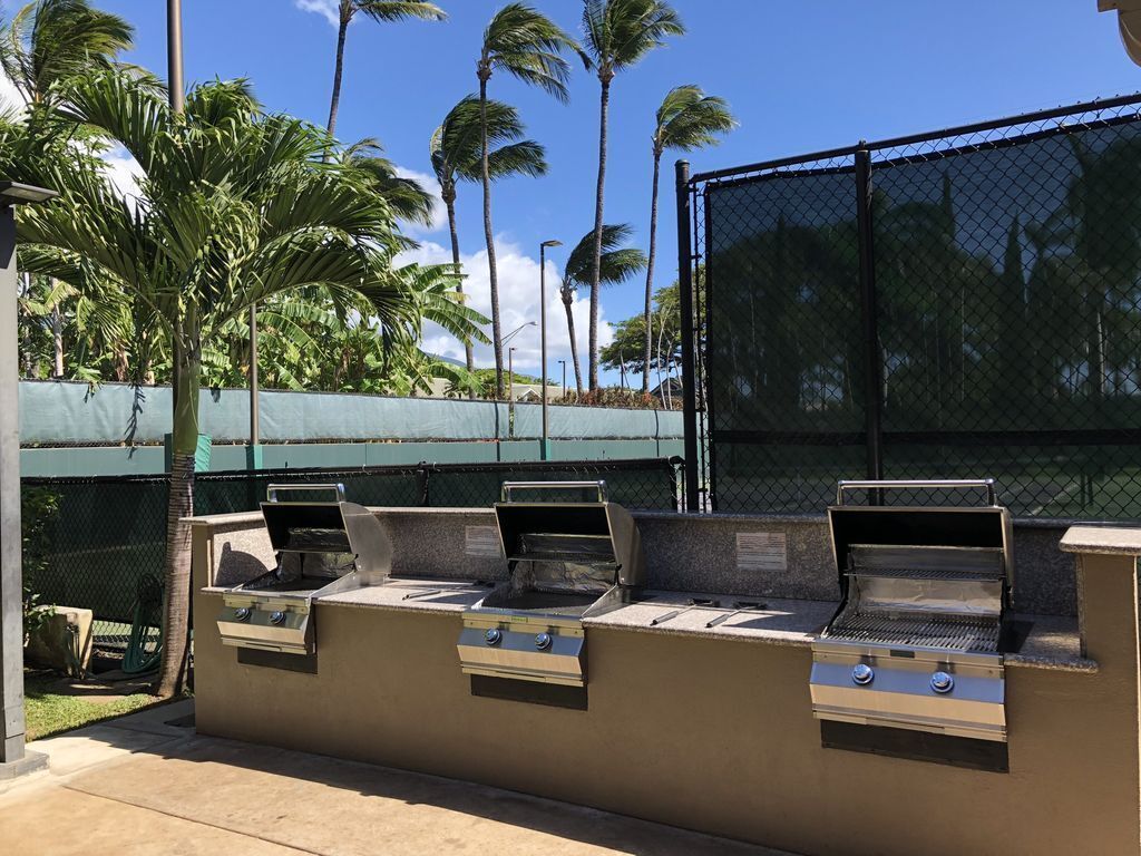 Ein Grillbereich mit drei Grills, umgeben von einem Maschendrahtzaun und Palmen.