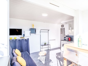 A modern living room with a grey sofa, a flat-screen TV, a small table, and a kitchen area with a microwave, toaster, and kettle.