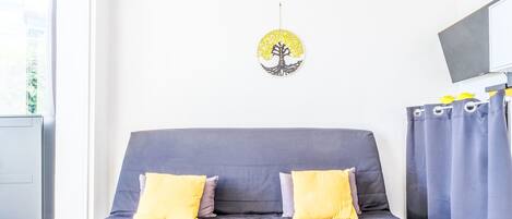 Ein modernes Wohnzimmer mit einem grauen Sofa, zwei gelben Kissen, einem Wand-Fernseher und einem Fenster mit Vorhängen.