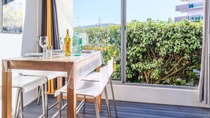 Un espace repas moderne avec une table dressée pour deux personnes, une bouteille de spiritueux et un verre. La pièce est dotée de grandes fenêtres qui offrent une vue sur un immeuble et sur la verdure.