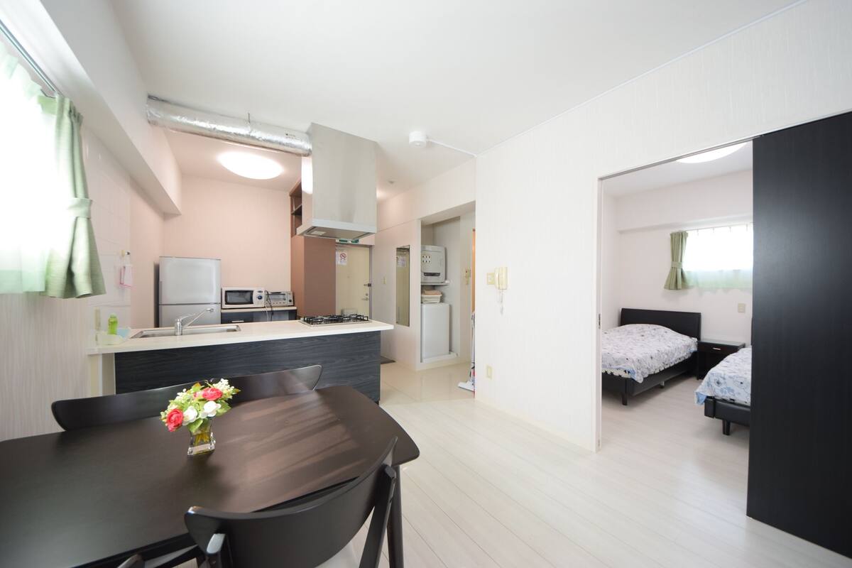 Een moderne keuken met een centraal eiland, een eethoek met tafel en stoelen, en een slaapkamer met een bed en nachtkastje.