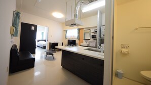 A modern kitchen with a central island, a dining area with a black sofa, and a visible bathroom with a toilet and sink. - SUMUKA Asato Station (Naha)