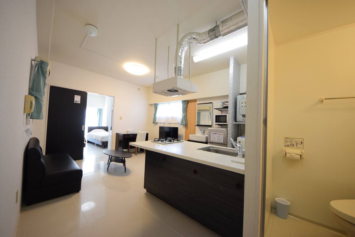 Een moderne keuken met een centraal eiland, een zithoek met een zwarte bank en een zichtbare badkamer met toilet en wastafel.