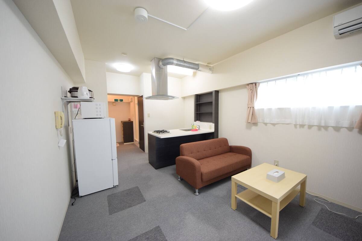 Een compacte woonruimte met een kitchenette, een bank, een tafeltje en een raam met gordijnen.