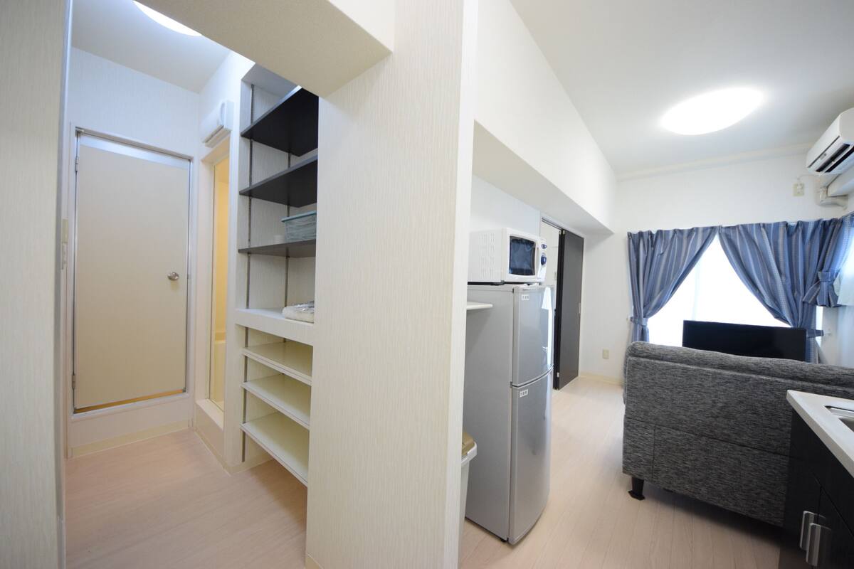 Een knus woongedeelte met een kitchenette, een grijze bank en een witte koelkast.