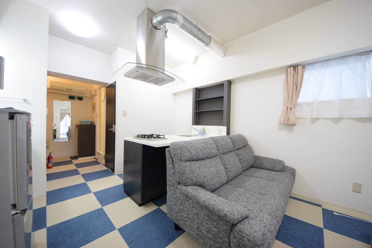 Een knus woongedeelte met een kitchenette, een bank, een zwart-wit geruit vloertje en een raam met gordijnen.