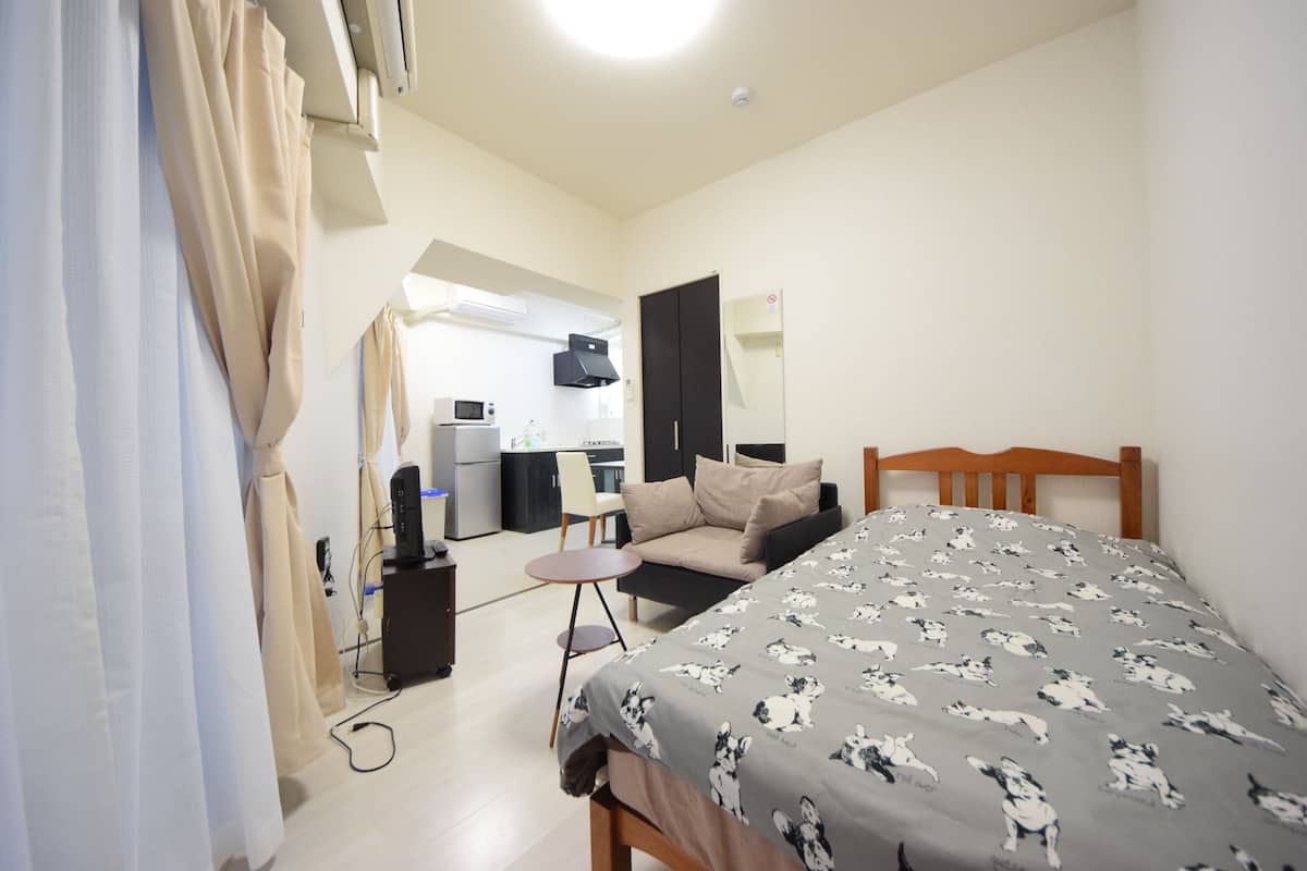 Een compacte hotelkamer met een bed, een klein tafeltje, een bank en een kitchenette op de achtergrond.