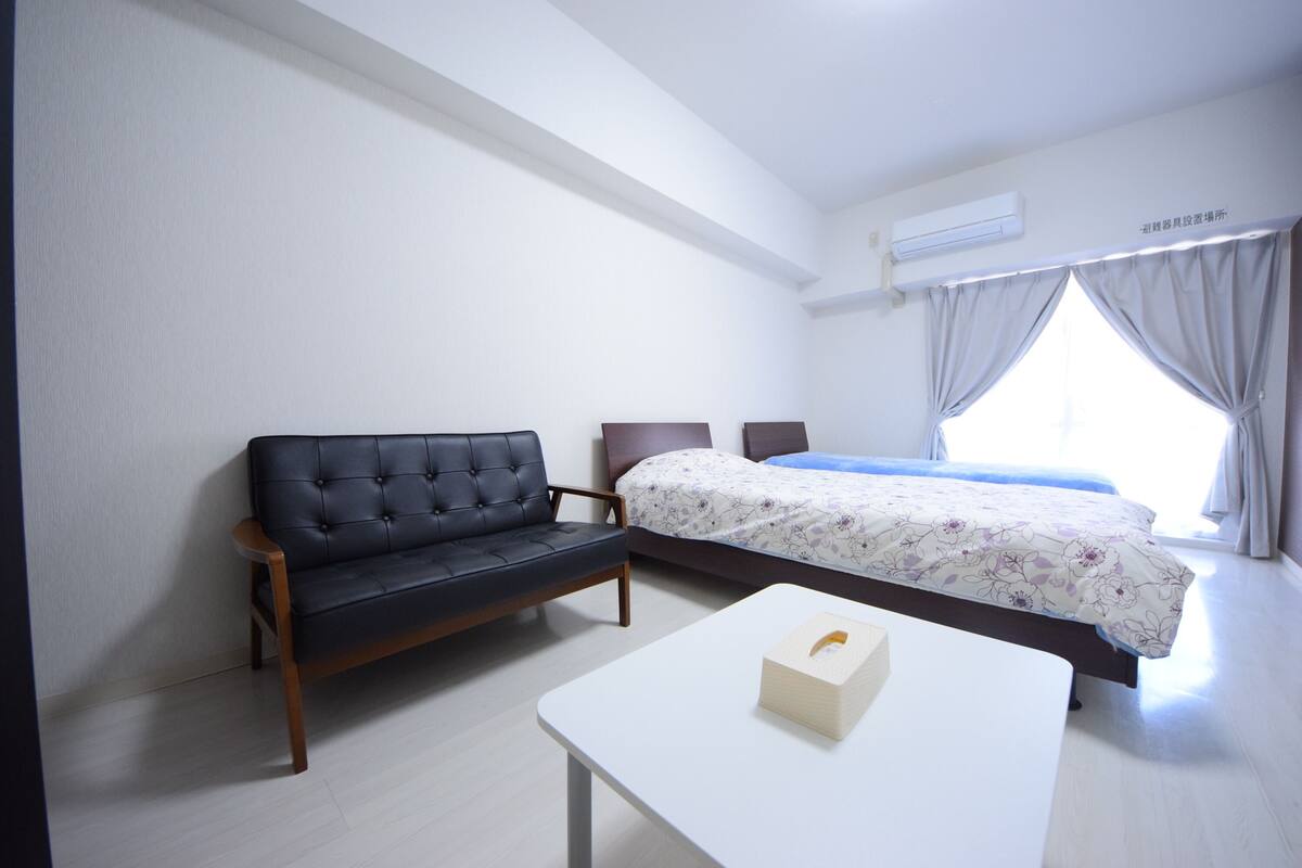 Een slaapkamer met een zwarte bank, een bed met bloemenbeddengoed, een witte nachtkastje en een raam met gordijnen.