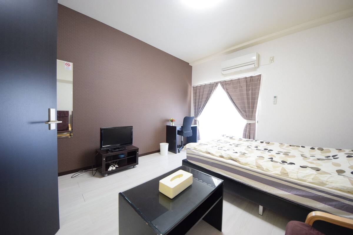 Hotelkamer met een bed, een televisie op een standaard, een klein tafeltje met zakdoekendoos, een raam met gordijnen, en een airconditioning aan de muur.