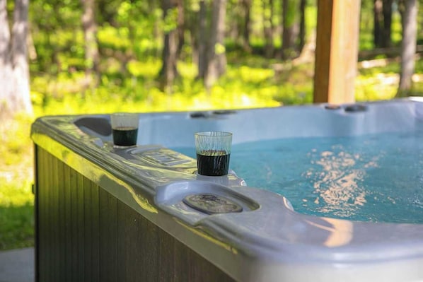 Un bain à remous avec deux verres remplis d’un liquide sombre, entouré d’une terrasse en bois et bordé d’une zone boisée.