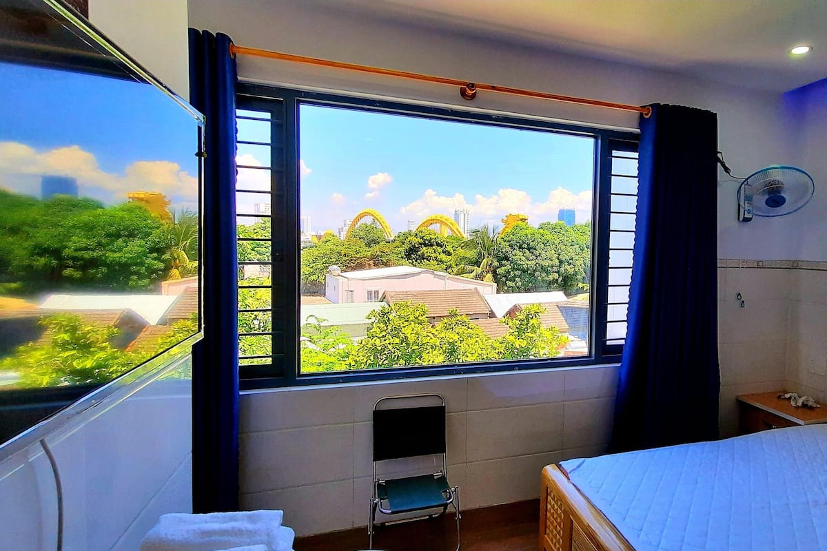 A hotel room with a large window, a bed, a chair, a bedside table, and a wall-mounted fan.
