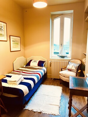 Een kleine, gezellige slaapkamer met een enkel bed, een stoel, een tafel en een raam met uitzicht op de zee.