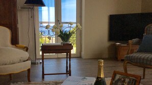 En stue med sofa, lænestol, et bord med bøger, en champagneflaske og et indrammet foto.