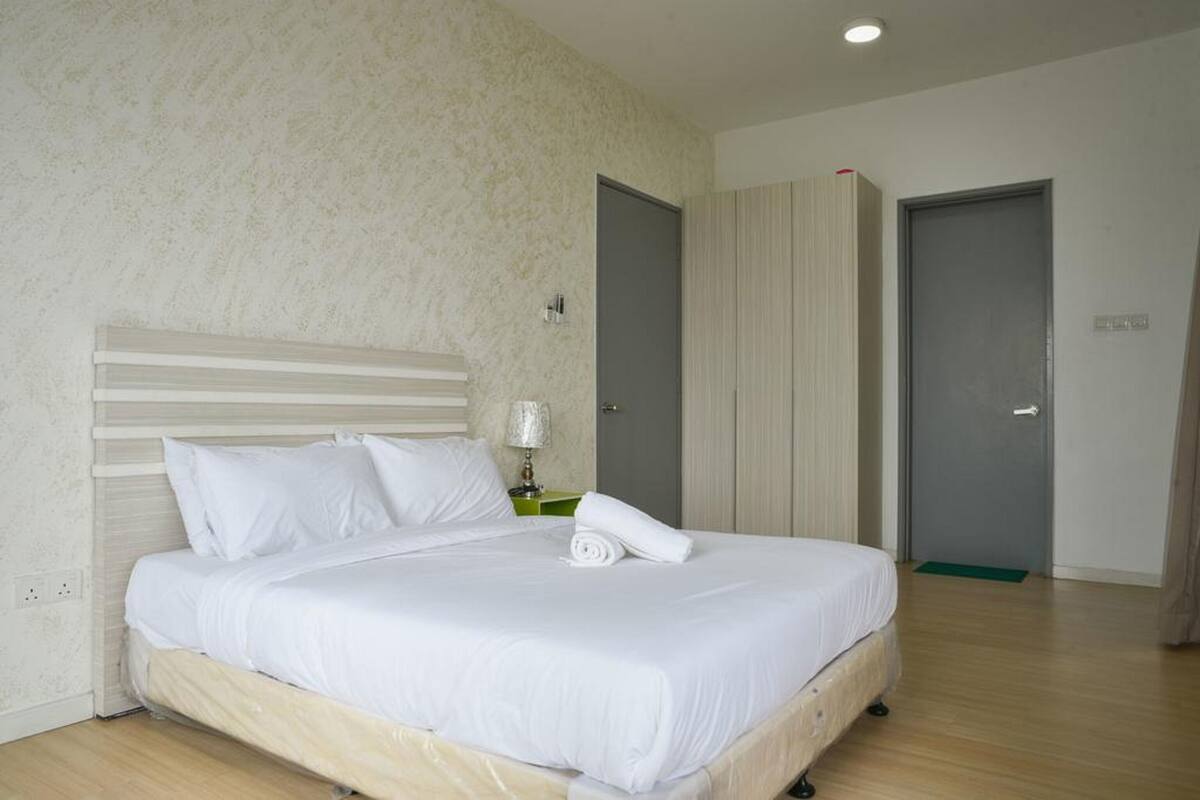 Un lit bien fait, avec du linge de lit blanc et une serviette enroulée, une lampe de chevet et une table de chevet verte, dans une pièce dont le mur est texturé et le sol en bois.